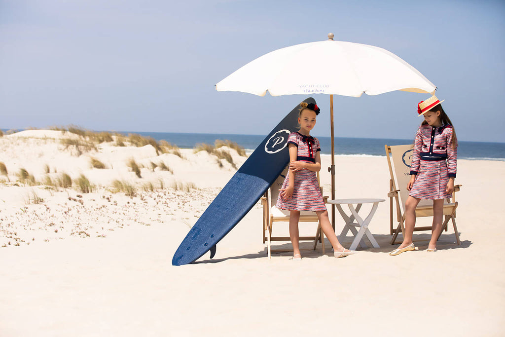 Girls in premium European-style dresses from Bambino Babe posing on a beach with surfboard and umbrella